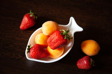 composition with a fruit plate against a dark wooden table. The fruit as a whole. Assorted apricots, strawberries iin a white deep plate in the shape of a fish. Vegetarian menu. raw food. healthy food
