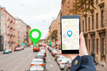 A woman's hand in a jacket holds a smartphone with an online map app. In the background is a blurred road with a traffic and icon of location.The concept of Internet navigation and modern technologies