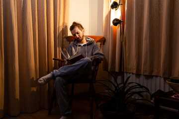 A 20 something man sitting in an arm chair reading by lamp light on a dark winter evening. Life and after dark concept