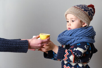 Sick child in clothes, toddler boy is standing on the grey background with a fever. llness child. Mother gives his son a lemon and a cup