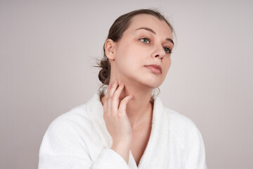 young woman in a white robe, touches her neck. Studio shot over light background. Facial care concept.