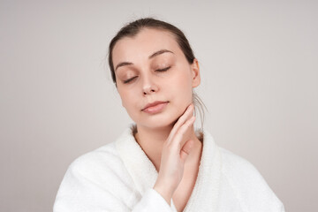 A pretty young woman in a white robe touches the delicate skin of her neck, closing her eyes. Studio shot over light background. Facial care concept.