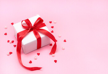 Gift box with a red ribbon bow, small sugar hearts and white beads on trendy pink background. Flat lay, top view Valentine's Day banner or greeting card.