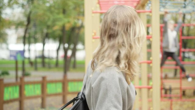 Happy beautiful slim Caucasian woman talking to blurred boy playing on playground at background, and turning to camera smiling. Portrait of charming young mother enjoying autumn weekend with son.