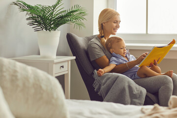 Happy young mommy reading first books to her little son sitting in armchair in cozy bedroom