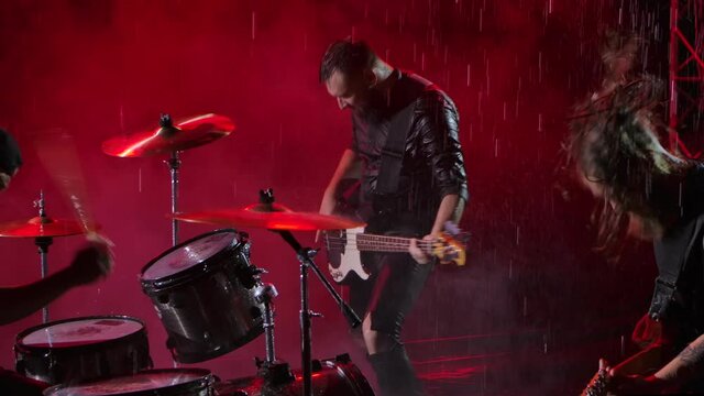 Camera slides over a group of rock musicians who play their instruments with crazy emotions in a dark studio illuminated by red lights. Drum set, electric and bass guitar. Close up. Slow motion.