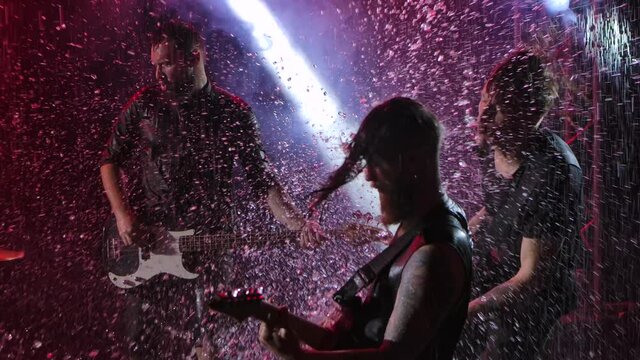 Live concert of rock band in dark studio illuminated by red light. Three guitarists with crazy emotions play instruments and wave their heads with long hair to the beat of music. Close up. Slow motion