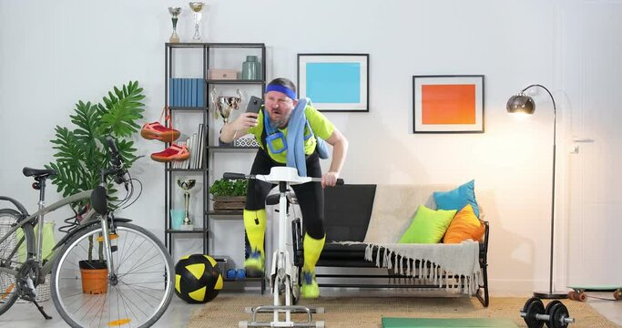 funny energetic athlete on exercise bike training at home and taking selfie , Sportsman in retro sportswear recording a cool cycling video on camera in an apartment.