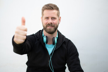Relaxed smiling sportsman sitting outdoors, wearing headphones with thumb up.