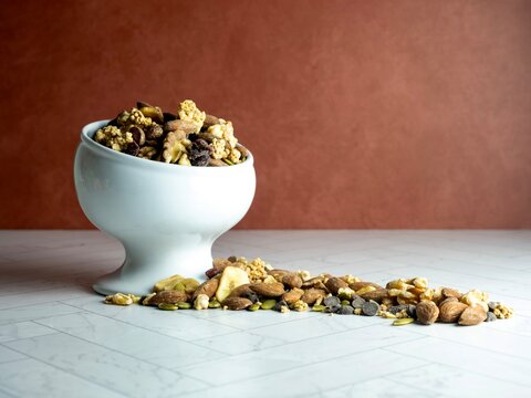 White raised bowl fill of trail mix, nuts and dried fruit spilling out onto the herringbone counter with a terra-cotta wall behind.  Peanuts, granola, pecans, walnuts, banana slices, cranberry.