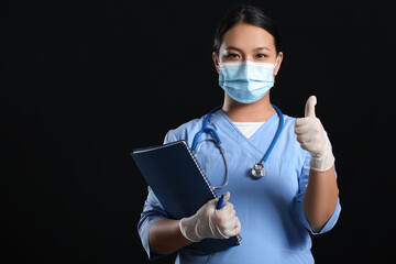 Portrait of female Asian doctor in medical mask showing thumb-up on dark background