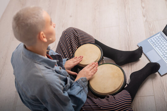 A young hipster woman sits on the floor at home and watches educational videos on mini bongs. Girl remotely learns to play traditional ethnic drums.