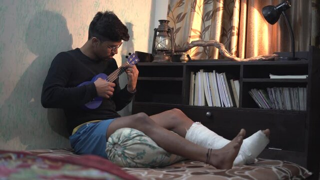 Young Indian man playing ukelele while sitting on the bed with a fractured leg.