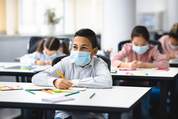 Boy in mask sitting at desk in classroom