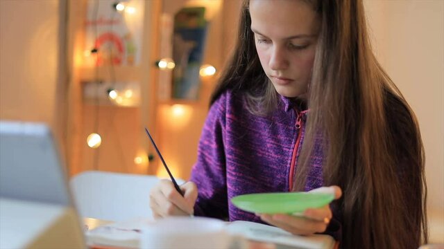 Young girl drawing with brush on paper and looking lesson on the tablet