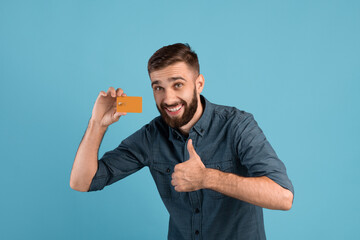 Cheerful young guy with credit card showing thumb up gesture on blue studio background