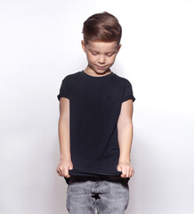 Cool little model boy wearing black blank t-shirt and jeans posing on gray studio background. Stylish schoolboy caucasian child looking down and adjusting clothes. Childhood style and fashion concept.