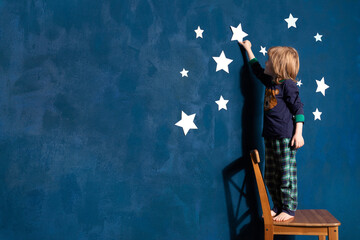 Little boy in pyjamas decorate blue bedroom wall with white paper stars. Dreaming curious caucasian...