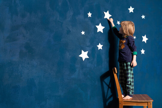 Little boy in pyjamas decorate blue bedroom wall with white paper stars. Dreaming curious caucasian preschool child in sleepwear touch star in children's room interior, copy space for store ad.