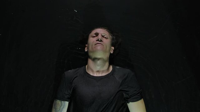 Top view of the leader of a rock band lies on the water surface in a dark studio. A man in black clothes lit by lights looks at the camera. Close up. Slow motion.