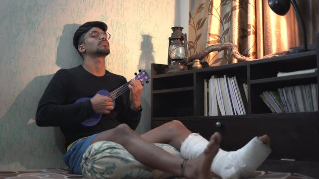 Young Indian man playing ukelele while sitting on the bed with a fractured leg.