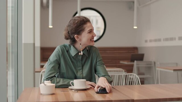 Medium shot of young curly-haired Caucasian woman sitting at cafe table, waiting for her boyfriend. Then young Caucasian man coming, sitting nearby and kissing her on cheek