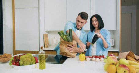 Attractive happy smiling married couple unpacking food paper bag full of different vegetables in the contemporary kitchen when came home from market
