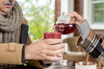 Hand of senior woman pouring herbal tea into ceramic mug held by her husband