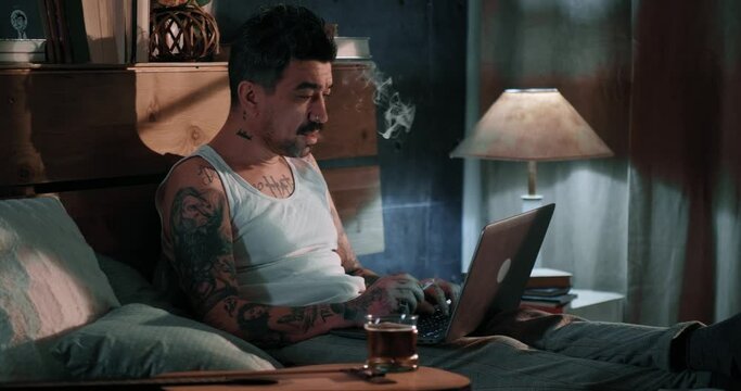 Tattooed man with a cigar in his undershirt uses a video call for work from home, relaxed in bed with a cigar and a beer. Different people, a different concept