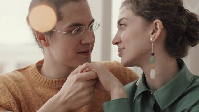 Close up of good-looking young Caucasian man wearing eyeglasses kissing hand of beautiful young woman wearing stylish earrings while looking in her eyes