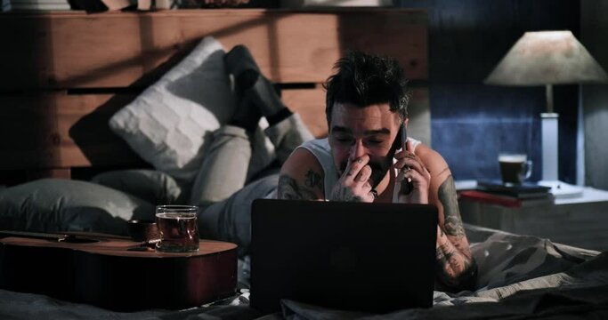 Musician chats with a friend using the phone. A tattooed man in an undershirt uses a laptop and talks with a smart device, drinking beer. Enjoying