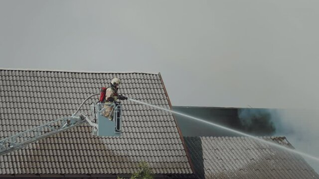 Shot of exhausted firefighter extinguishing a large building caught on fire standing on a ladder truck. A lot of smoke while he is hosting down hot spots.