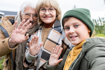 Cheerful boy and his affectionate grandparents waving hands and making selfie
