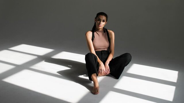 a girl sits on a light figure from the window in the studio on a white background, dressed in black trousers and no shoes with bare legs