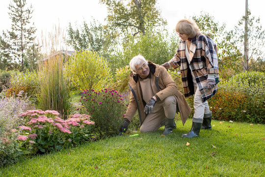 Retired Female In Eyeglasses And Warm Coat Helping Her Husband In The Garden