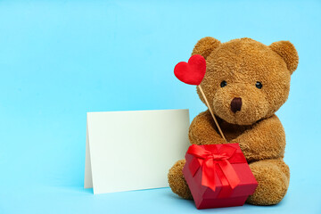 Cute teddy bear with red heart, gift box and blank card on light blue background. Valentine's day celebration