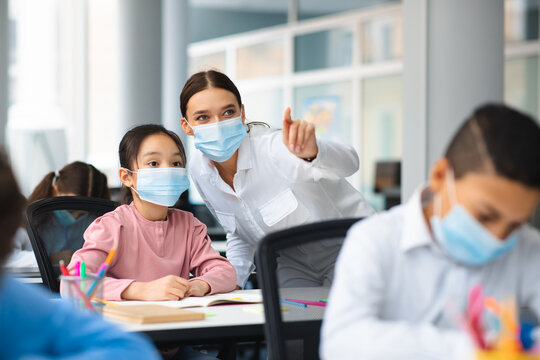 Female teacher in disposable face mask helping small asian girl