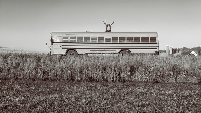 Wide angle shot of a man standing on the top of a bus in the middle of the countryside and raising his arms to the sky. Black and white.