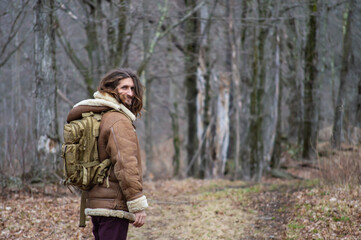 Fototapeta na wymiar Rear view of a young man with long hair, sheepskin winter coat and green military backpack looking at camera while walking in the woodlands.