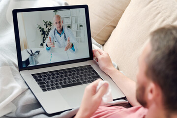 Young sick man with laptop and crumpled handkerchief consulting online doctor