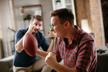 Two young friends enjoying at home. Men drinking beer and watching sports game on tv..