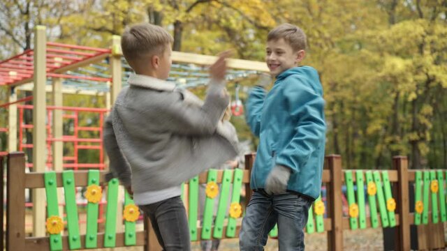 Joyful Caucasian boys sharing secrets and giving high-five standing on playground in autumn park with blurred mothers at background. Happy relaxed friends whispering on ear and laughing outdoors.
