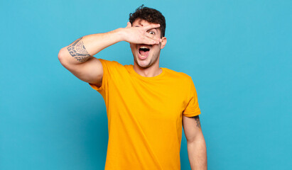young man looking shocked, scared or terrified, covering face with hand and peeking between fingers