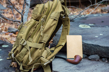 Close up advertising style shot of a green army tactical backpack, a paper notebook and a vintage pipe disposed on rock steps in the woodlands.