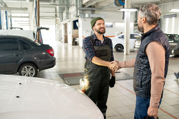Happy young bearded technician of car service shaking hand of male client