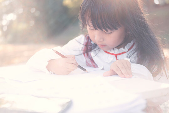 asian little girl lying drawing or made homework in the paper book for preschool kids in the home garden park with sun ligh flare