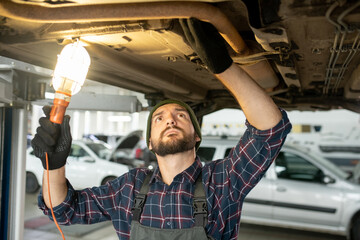 Young repairman in gloves and uniform holding lamp while checking loosen details