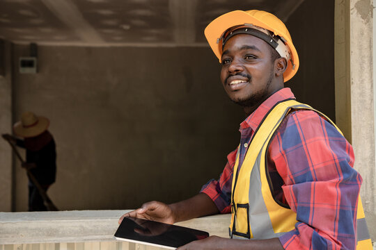 Africa American Foreman Controls Home Building And Plans Projects By Tablet To Invest In Real Estate On The Job Site. Back View Of House Property With Laborer.