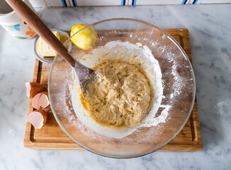Glass bowl full of cake mixture, and ingredients on the side to make cake, sponge biscuits. On wooden board in kitchen