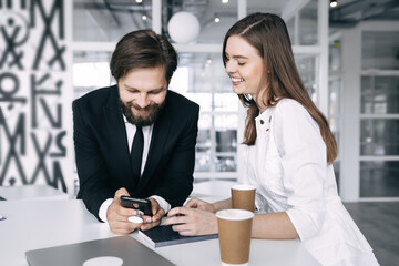 cheerful girl and bearded man team of startups with smartphone drinks coffee in the office